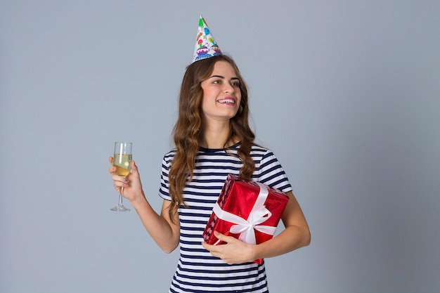 Młoda uśmiechnięta kobieta w świątecznej czapce trzymająca czerwony prezent i kieliszek szampana