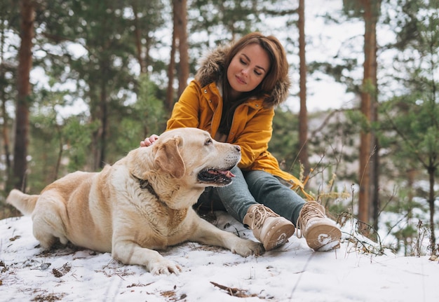 Młoda uśmiechnięta kobieta w żółtej kurtce z dużym miłym białym psem labradorem spacerującym w zimowym lesie