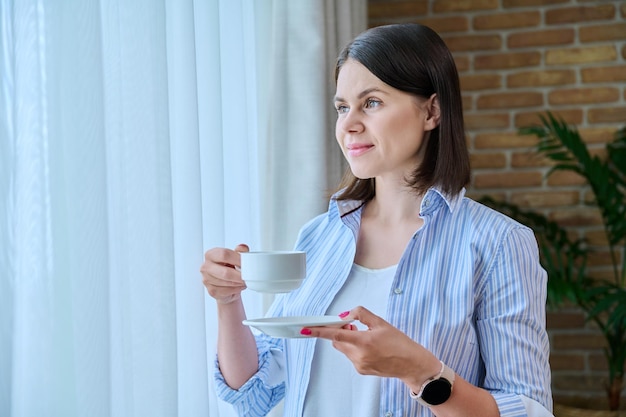 Młoda uśmiechnięta kobieta z filiżanką porannej kawy w domu przy oknie