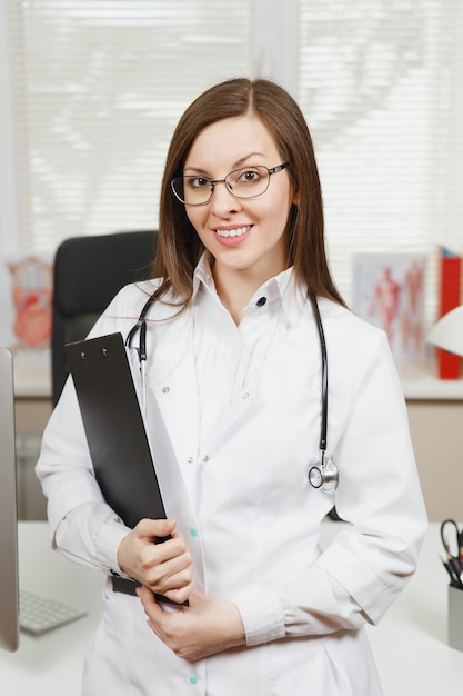 Młoda uśmiechnięta lekarka stojąca przy biurku ze schowkiem i dokumentami medycznymi w jasnym biurze w szpitalu