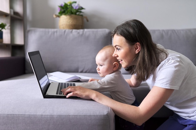 Młoda uśmiechnięta mama z małym dzieckiem patrzą razem na monitor laptopa siedząc w domu na kanapie