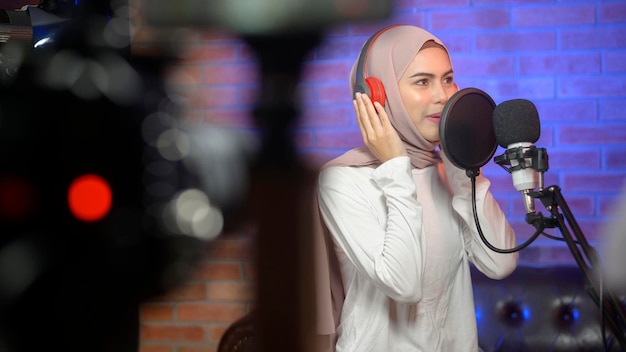 Młoda uśmiechnięta muzułmańska piosenkarka w słuchawkach z mikrofonem podczas nagrywania piosenki w studiu muzycznym z kolorowymi światłami.