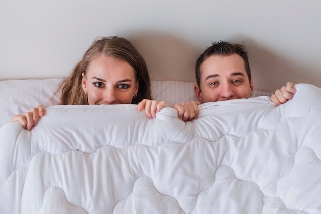 Zdjęcie młoda uśmiechnięta para leżąc na łóżku i zaglądając z kołdrą