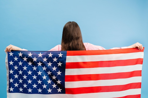 Młoda uśmiechnięta szczęśliwa kobieta nosi zwykłe ubrania trzymając amerykańską flagę i patrząc na kamerę odizolowaną na niebieskim tle