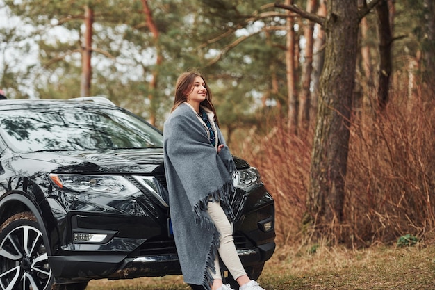 Młoda wesoła dziewczyna stojąca w pobliżu nowoczesnego czarnego samochodu na zewnątrz w lesie