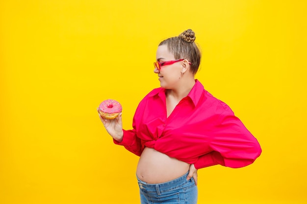Młoda wesoła kobieta w ciąży w różowej koszuli izolowana na żółtym tle trzymająca pączki z wesołym wyrazem twarzy Słodkie jedzenie podczas ciąży