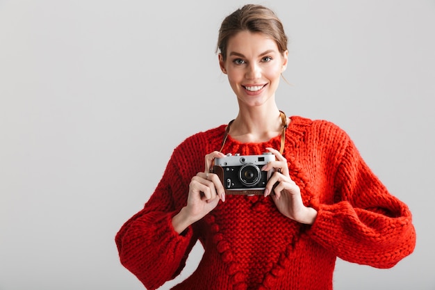 młoda wesoła kobieta w czerwonym swetrze uśmiecha się i używa retro kamery odizolowanej na białym tle