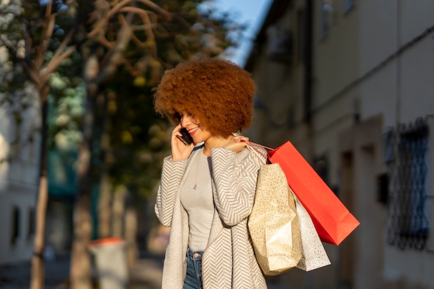 Młoda wesoła kobieta z afro włosami, trzymając torby na zakupy i rozmawiając na smartfonie na ulicy miasta