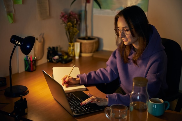 Młoda wesoła, pewna siebie i szczęśliwa freelancerka pracuje wieczorem na laptopie w domu