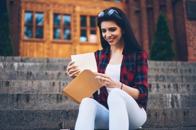 Młoda wesoła studentka czyta list z papierowej paczki podczas siedzenia w wiosennej scenerii miejskiej