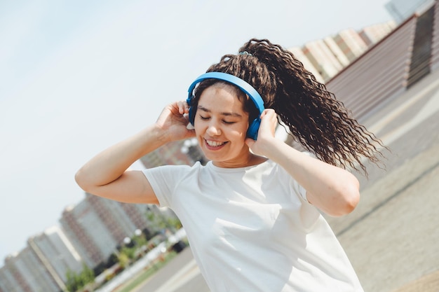 Młoda wesoła szczęśliwa kobieta z dredami ubrana w białą koszulkę tańczy słuchając muzyki ze słuchawkami odpoczywając w parku miejskim spacerując aleją Koncepcja miejskiego stylu życia