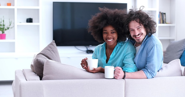 młoda wieloetniczna para siedzi na kanapie w domu, pijąc kawę, rozmawiając, uśmiechając się.