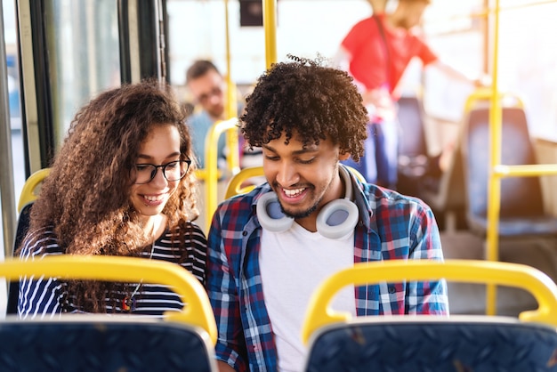 Młoda wielokulturowa para patrzeje w dół i ono uśmiecha się podczas gdy siedzący i jadący w miasto autobusie.