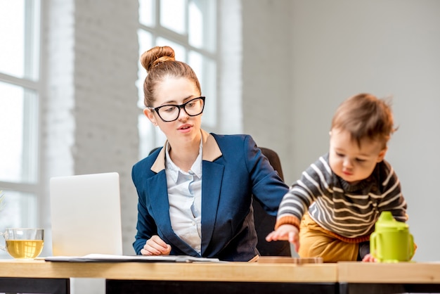 Zdjęcie młoda wielozadaniowa biznesmanka ubrana w garnitur, pracująca z laptopem i dokumentami siedząca z synkiem w biurze