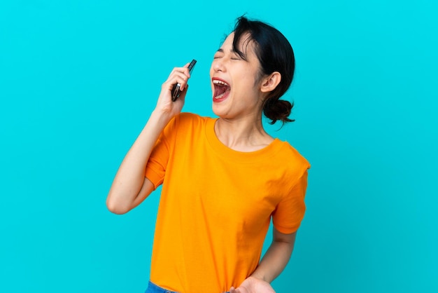 Młoda Wietnamka odizolowana na niebieskim tle, korzystająca z telefonu komórkowego i śpiewająca