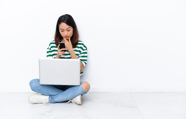 Młoda Wietnamka z laptopem siedząca na podłodze na białej ścianie, myśląca i wysyłająca wiadomość
