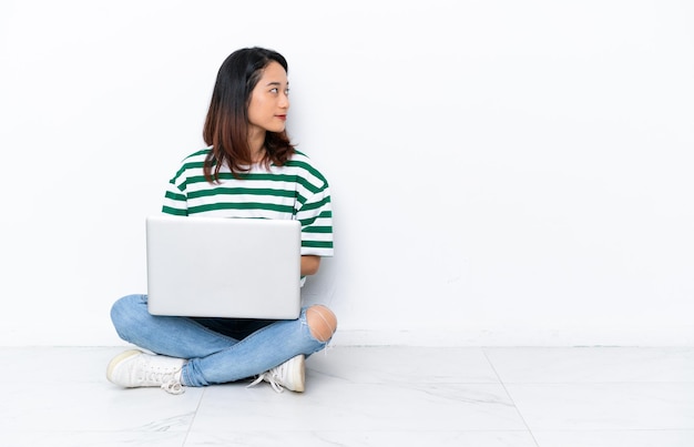 Młoda Wietnamka z laptopem siedząca na podłodze na białej ścianie w pozycji bocznej
