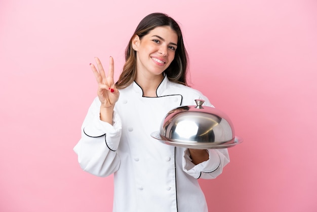 Młoda włoska kobieta kucharz trzyma tacę z pokrywką na białym tle na różowym tle szczęśliwa i licząca trzy palcami