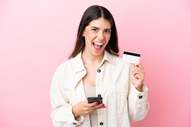 Młoda Włoszka odizolowana na różowym tle, kupująca za pomocą telefonu komórkowego i trzymająca kartę kredytową ze zdziwionym wyrazem twarzy