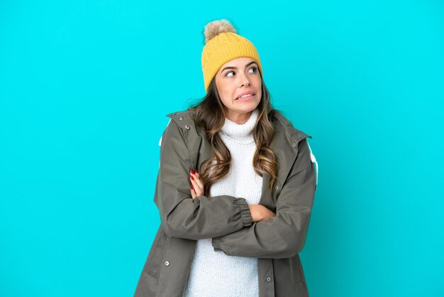 Młoda Włoszka ubrana w zimową kurtkę i czapkę na białym tle na niebieskim tle, wykonująca gest wątpliwości podczas podnoszenia ramion