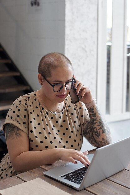 Młoda współczesna bizneswoman w casualwear i okularach mówiąca przez telefon komórkowy i patrząca na ekran laptopa