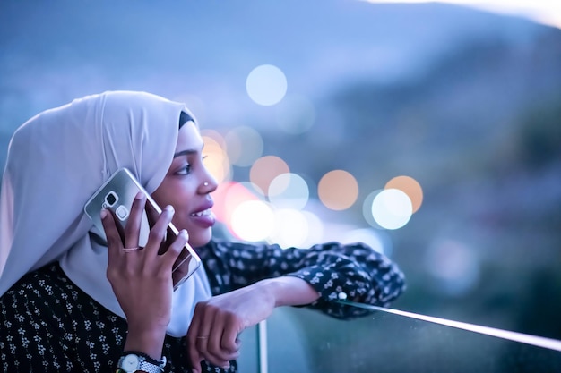 Młoda współczesna muzułmanka ubrana w szalik na ulicy miasta w nocy, SMS-y na smartfonie ze światłem miasta bokeh w tle