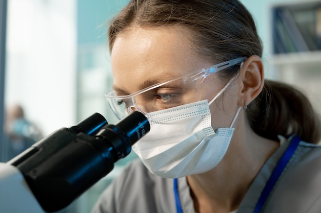 Młoda współczesna naukowiec w masce ochronnej i okularach, patrząc na próbki chemiczne lub biologiczne w mikroskopie w laboratorium