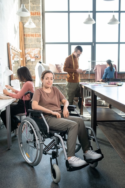 Młoda wyłącza krawcowa w stroju codziennym siedząca na wózku inwalidzkim przed kamerą przeciwko kolegom pracującym nad nową kolekcją mody