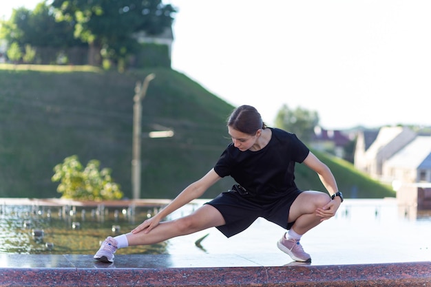 Młoda, wysportowana i wysportowana dziewczyna w czarnych ubraniach rozciągająca się po treningu w pobliżu publicznej fontanny w miejskim parku miejskim Fitness, miejskie jogging i koncepcja zdrowego stylu życia