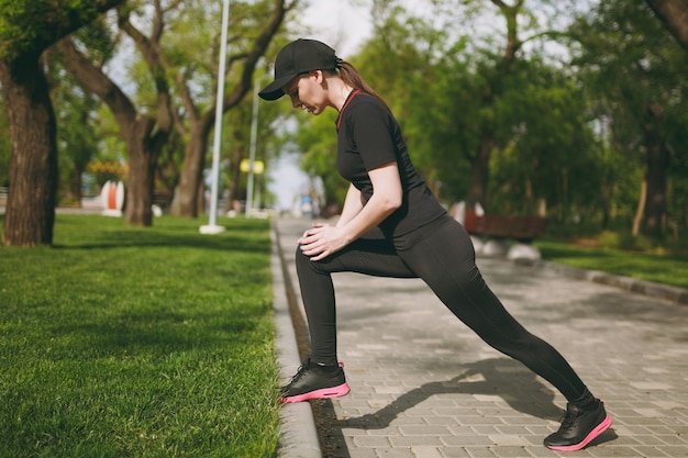 Młoda Wysportowana Piękna Brunetka W Czarnym Mundurze, Czapka Robi ćwiczenia Rozciągające Sportowe, Rozgrzewka Przed Bieganiem Lub Treningiem, Stojąc W Parku Miejskim Na Zewnątrz