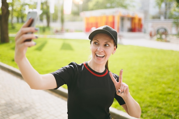 Młoda wysportowana uśmiechnięta piękna brunetka dziewczyna w czarnym mundurze i czapce robi selfie na telefonie komórkowym podczas treningu, pokazując znak zwycięstwa, stojąc w parku miejskim na zewnątrz