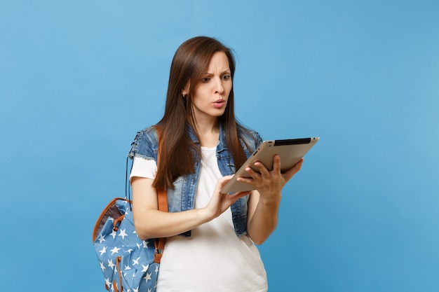 Młoda Zaniepokojona Studentka W T-shirt, Dżinsowe Ubrania Z Plecakiem, Przy Użyciu Pracy Na Komputerze Typu Tablet Pc Na Białym Tle Na Niebieskim Tle. Edukacja Na Uniwersytecie. Skopiuj Miejsce Na Reklamę.