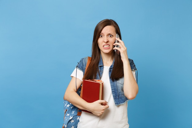 Młoda zaniepokojona winna studentka z plecakiem trzymająca szkolną książkę rozmawiającą na telefonie komórkowym prowadzącą rozmowę na białym tle na niebieskim tle. Edukacja w koncepcji liceum uniwersyteckiego.