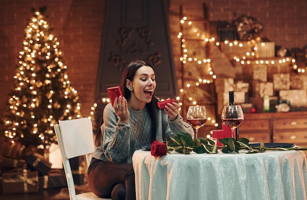 Młoda zaskoczona brunetka siedzi przy stole w restauracji i trzyma czerwone pudełko z prezentem