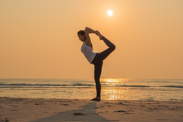Młoda zdrowa kobieta ćwiczy joga pozę na plaży przy wschodem słońca.