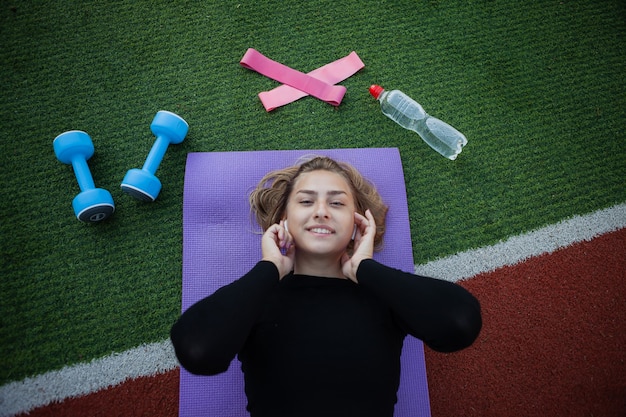 Młoda zdrowa sprawna kobieta odpoczywa leżąc na macie i listem muzyki. Trening na zewnątrz. Fitness, koncepcja sportu. Zdrowy tryb życia