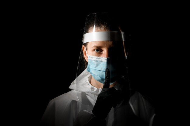 Młoda żeńska pielęgniarka w medycznej masce i ochronnej tarczy pokazuje gest ciszy.