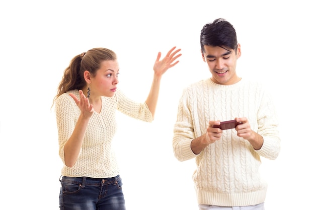 Młoda zła kobieta z kasztanowym kucykiem kłócąca się z młodym uśmiechniętym mężczyzną, który używa smartfona