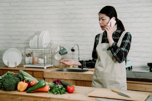Młoda zły smutna azjatycka chińska kobieta gospodyni domowa rozmowa na telefon komórkowy w jasnej, nowoczesnej kuchni. wiele zielonych warzyw na stole licznika. szalona pani w fartuchu kłóci się online na telefonie komórkowym podczas gotowania