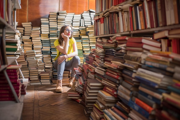 Młoda zmęczona studentka siedząca na stosie książek w starej bibliotece