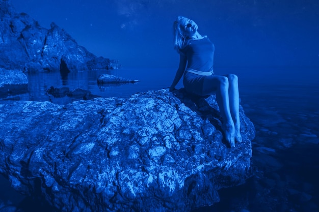 Młoda zrelaksowana kaukaska kobieta siedzi na kamieniu i cieszy się letnią nocą na tle rozgwieżdżonego nieba podczas długo wyczekiwanych wakacji w letnią noc. Koncepcja odpoczynku i regeneracji