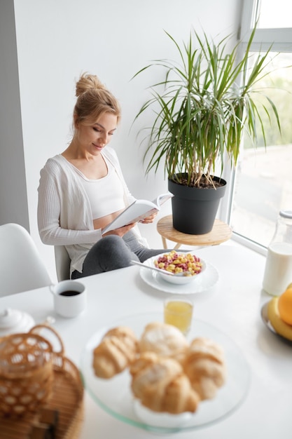 Młoda zrelaksowana kobieta czytająca po siedzeniu przy kuchennym stole po zdrowym śniadaniu w domu rano