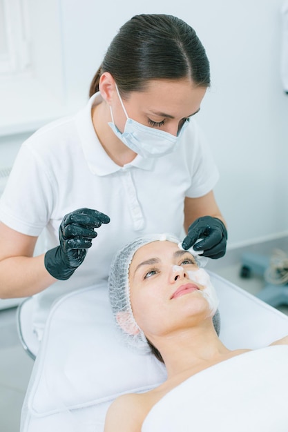 Młoda zrelaksowana kobieta leżąca na kanapie medycznej, podczas gdy kosmetolog w gumowych rękawiczkach myje jej twarz
