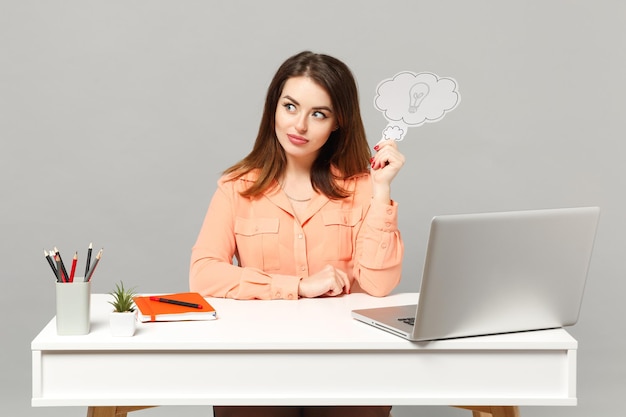 Młoda zszokowana kobieta w pastelowych ubraniach trzymająca powiedz chmurę z żarówką, siedzieć przy biurku z laptopem pc na białym tle na szarym tle. Koncepcja stylu życia kariery kariery osiągnięcia. Makieta miejsca na kopię.