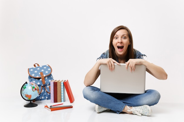 Młoda zszokowana studentka z otwartymi ustami, trzymająca się za laptop PC, siedząca w pobliżu globu plecaka szkolne książki na białym tle