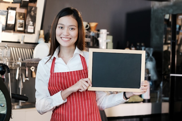 Młode azjatykcie kobiety Barista mienia chalkboard pusty menu z uśmiechniętą twarzą przy kawiarnia kontuaru tłem