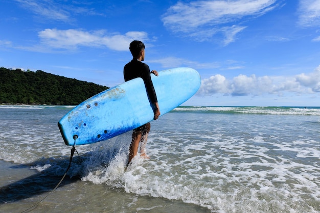 Zdjęcie młode dopasowanie i jędrne ciało nastoletniego faceta idącego do morza trzymającego miękką deskę gotową do sportów ekstremalnych surfowania na wakacjach na plaży w pogodny dzień błękitne niebo