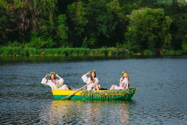 Młode dziewczyny w strojach ludowych pływają łodzią ozdobioną liśćmi i porostami. Słowiańskie święto Iwana Kupały.