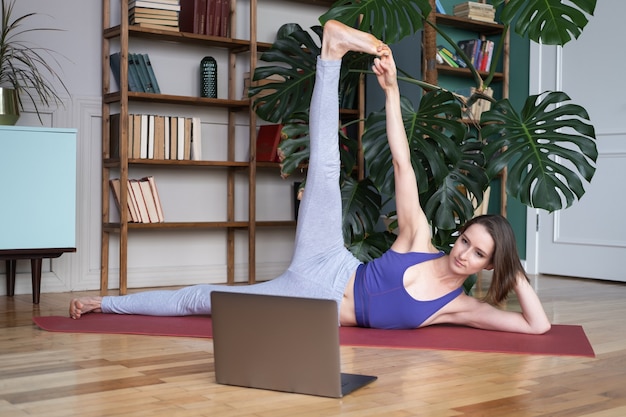 Młode kobiety praktykujących jogę w domu na macie do jogi, oglądając filmy na laptopie