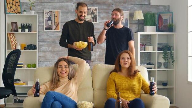 Młode kobiety siedzące na kanapie uśmiechnięte podczas oglądania telewizji, podczas gdy chłopcy jedzą frytki i dobrze się bawią.